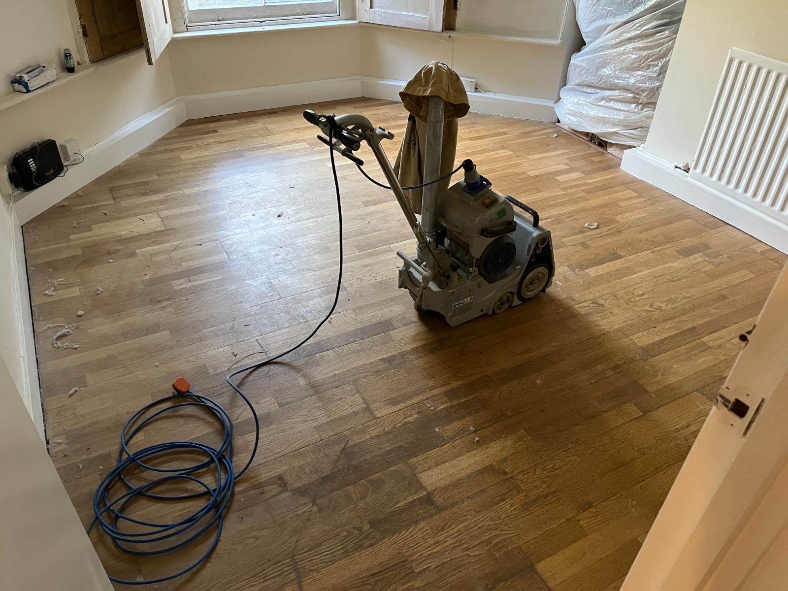 Professional Wood Floor Sanding machine standing in the centre of a room, working on an oak strip hardwood floor that shows contrasting sanded and unsanded areas.