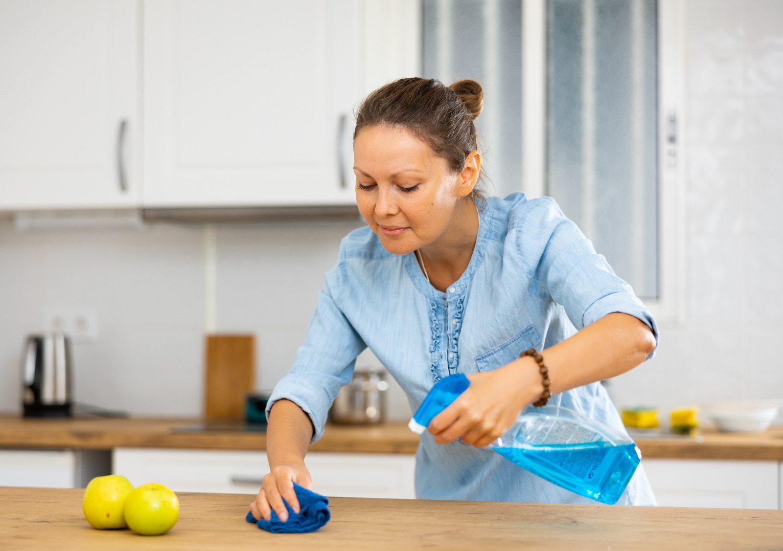 Worktop Refinishing aftercare – woman cleaning an oak wooden kitchen worktop with spray and cloth, keeping the restored surface and matching oak hardwood floor in top condition.
