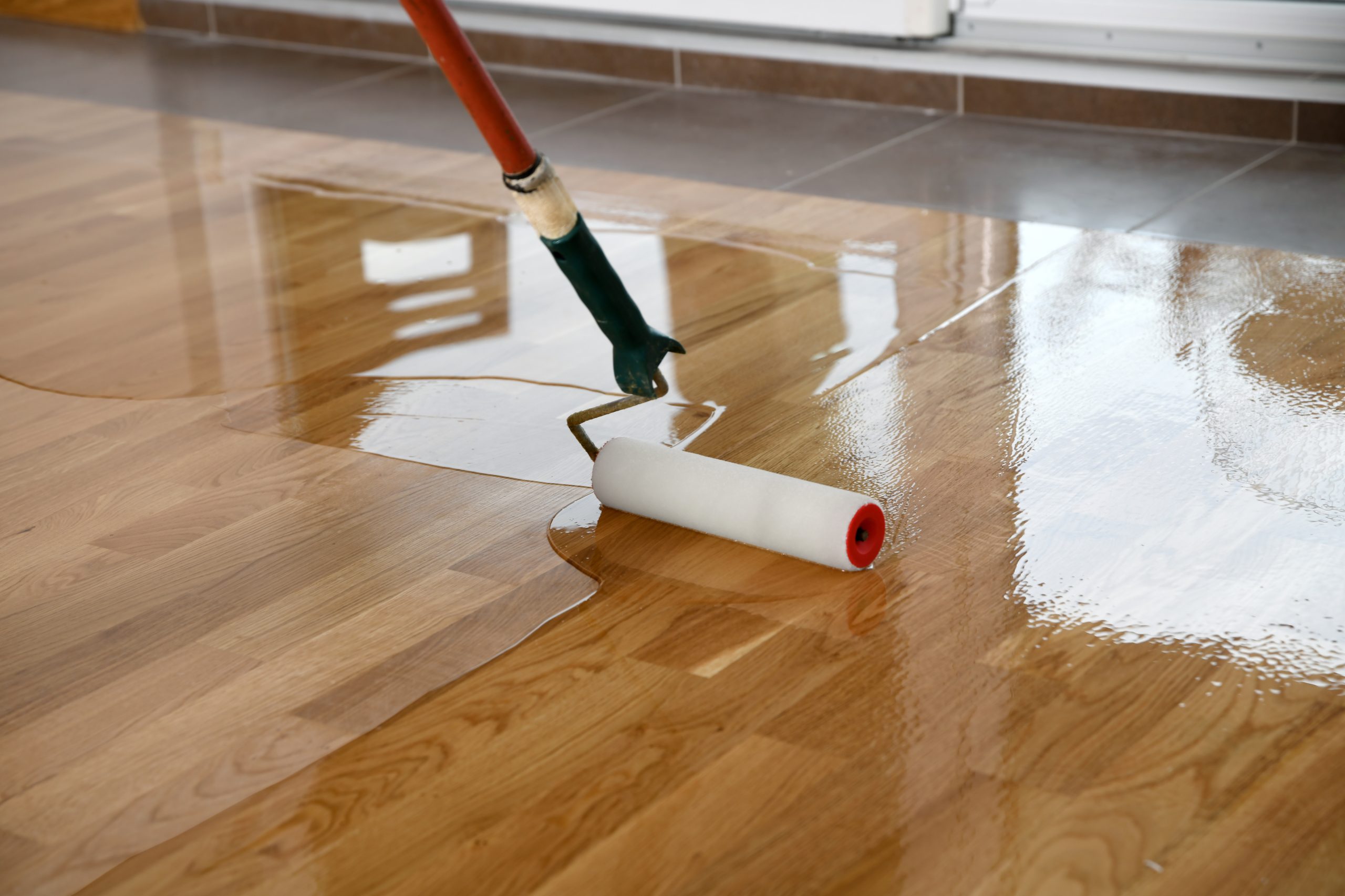 Hardwood Floor Refinishing in progress as a roller applies clear protective lacquer to an oak hardwood plank floor for a smooth, glossy finish.